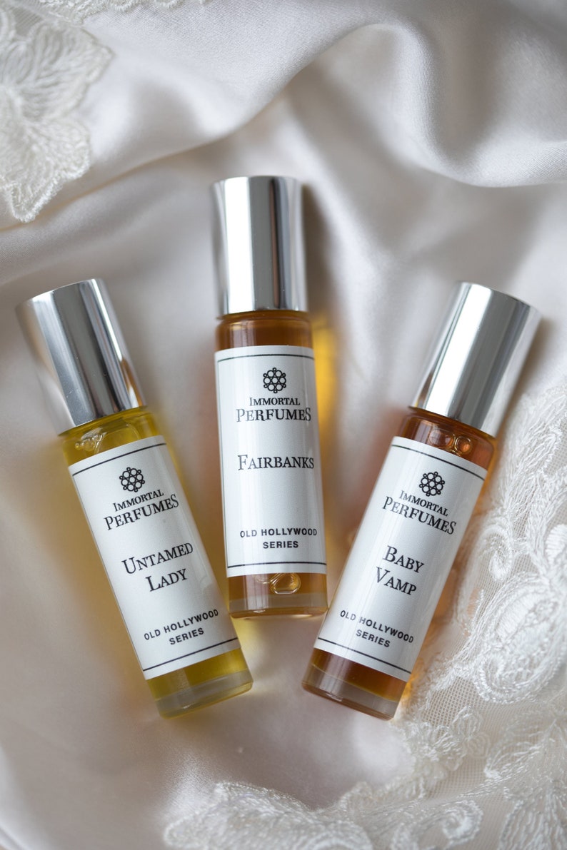 May include: Three small glass bottles of perfume with silver caps. The bottles are labeled with the text "Immortal Perfumes" and the names of the scents: "Fairbanks", "Untamed Lady", and "Baby Vamp". Each bottle is labeled with "Old Hollywood Series".