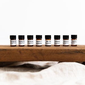 May include: Ten small brown glass bottles with black caps and white labels. The labels have the names of different perfumes on them, including "Georgiana", "Sharma B", "Bad Write", "Maitre d Immortial", "Mr. Ro Immortial", "Untame Immortial", "Artemis Immortial", and "Pemberton Immortial". The bottles are arranged in a row on a wooden surface.