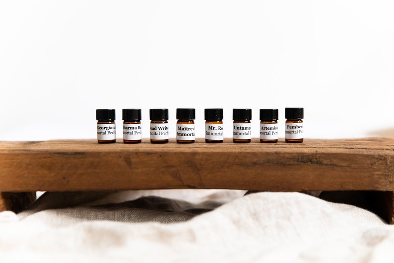 May include: Ten small brown glass bottles with black caps and white labels. The labels have the names of different perfumes on them, including "Georgiana", "Sharma B", "Bad Write", "Maitre d Immortial", "Mr. Ro Immortial", "Untame Immortial", "Artemis Immortial", and "Pemberton Immortial". The bottles are arranged in a row on a wooden surface.