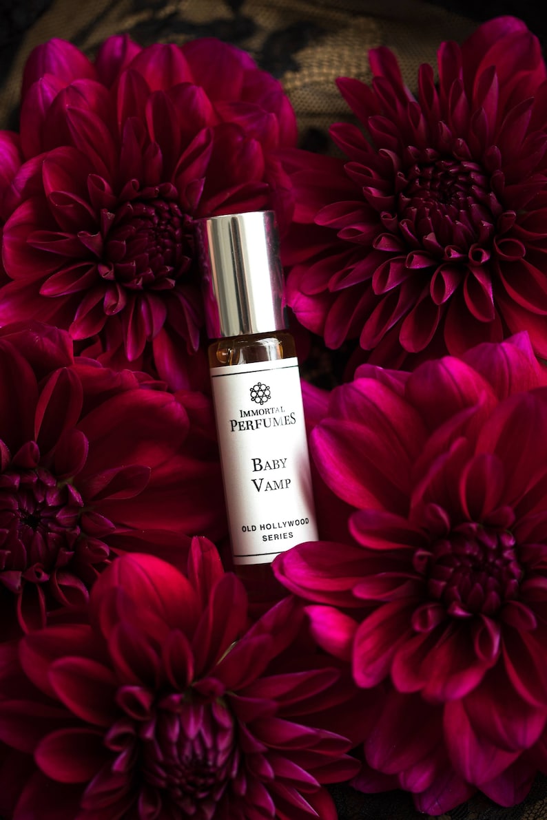 May include: A small glass bottle of perfume with a silver cap. The bottle is surrounded by deep red flowers. The label reads "Immortal Perfumes, Baby Vamp, Old Hollywood Series".