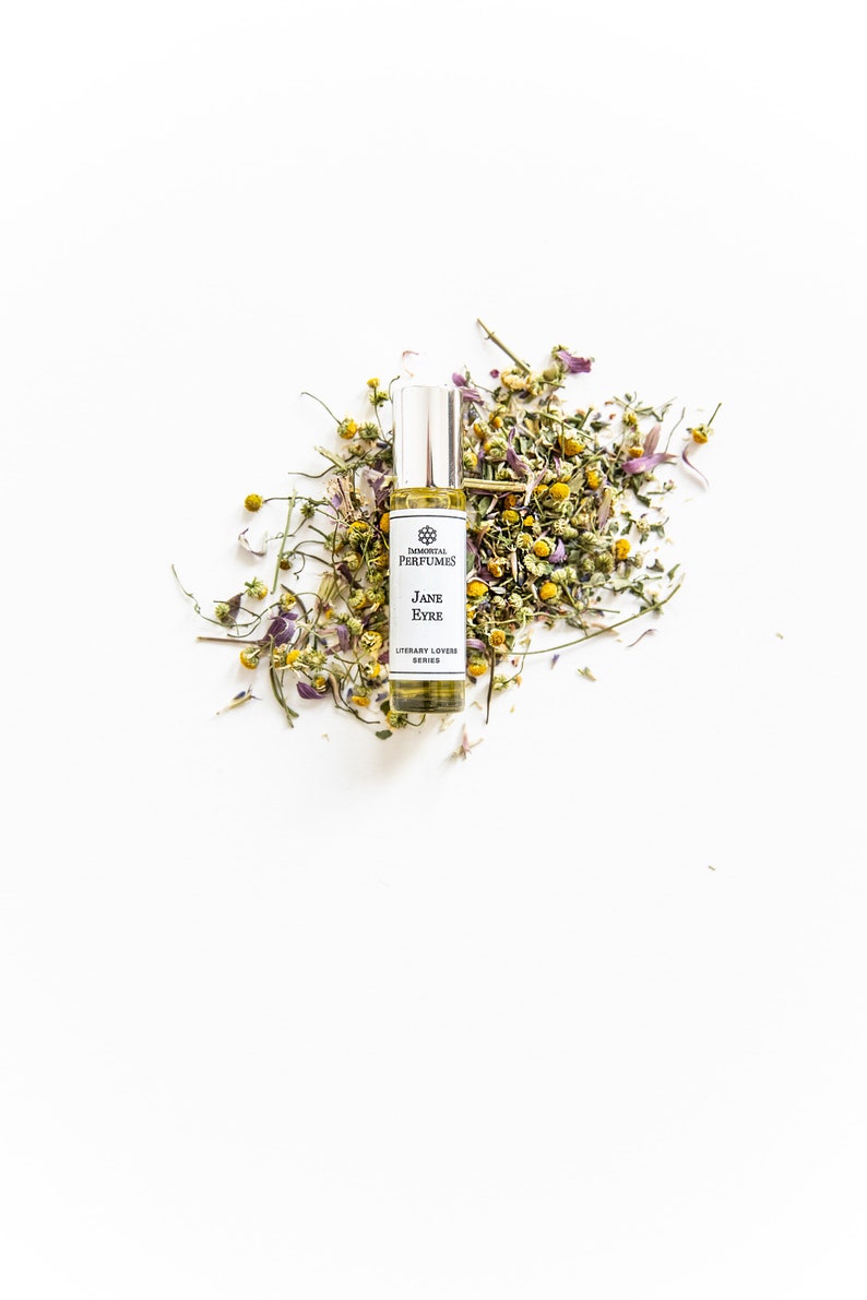 May include: A small glass bottle of perfume with a white label that reads "Elemental Perfumes Jane Eyre Literary Lovers Series". The bottle is surrounded by dried chamomile flowers.