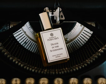 May include: A small glass bottle of perfume with a gold cap and a white label. The label reads "Immortal Perfumes, Death in the Afternoon, Dead Writers Series". The bottle is sitting on a vintage typewriter.
