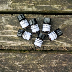 Puede incluir: Seis pequeños frascos de vidrio negro con etiquetas blancas que contienen aceite de perfume. Las etiquetas tienen los nombres "VIII", "Georgiana", "Lady Day", "Antoinette", "Archibald" y "Sharma Bush" impresos en ellas. Cada etiqueta también incluye el texto "Perfume Inmortal".