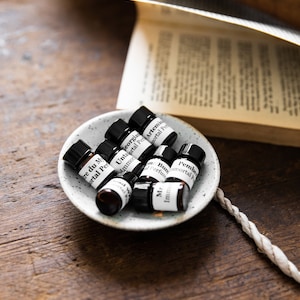 May include: A small white ceramic dish containing eight small glass bottles of essential oils. The bottles are labelled with the names of the scents, including "Tre du My", "Georgian", "Untaortal", "Artemis", "Pember", "Mr", "Imm", and "Bun".