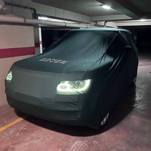 以下が含まれることがあります： 車両を覆うダークグリーンのカーカバー。ボンネット部分には白い文字でRANGE ROVERとプリントされています。カバーはフロントエンドにぴったりとフィットしており、その下にある車の明るい白色LEDヘッドライトが点灯しているのが見えます。