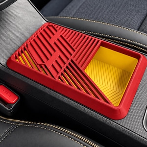 Op de afbeelding: Een rechthoekige rode en gele auto middenconsole organizer tray met een geometrisch lattenontwerp en een diep open opbergvak. De tray is geplaatst in het gestructureerde zwarte kunststof consolegebied van een voertuiginterieur.