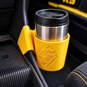 May include: A bright yellow plastic cup holder attachment mounted to a car center console, featuring a geometric embossed logo on the front. A stainless steel travel mug with a black lid is securely placed inside the yellow cup holder.