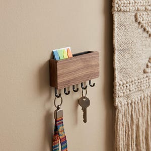 May include: A rectangular wall-mounted organizer made of brown wood with a top slot containing four colorful sticky notes. The unit features five black metal hooks along the bottom edge, with one silver key and a patterned fabric lanyard hanging.