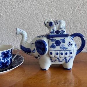 Puede incluir: Una tetera de cerámica blanca con forma de elefante, decorada con motivos florales azules y una pequeña figura de elefante en la tapa. A su lado hay una taza y un plato a juego, ambos con diseños florales azules sobre fondo blanco.