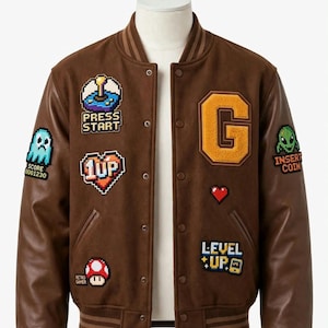 May include: A brown varsity jacket featuring pixelated patches including a joystick with the text PRESS START, a heart with 1UP, a mushroom with RETRO GAMER, a ghost with SCORE 0001230, a large letter G, a small heart, and a sign with INSERT COIN and LEVEL UP.