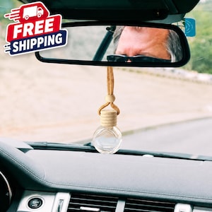 May include: A small, clear, spherical glass bottle with a light-colored wooden cap and a matching wooden bead, suspended by a tan cord from a vehicle's rearview mirror. The bottle is empty and features a ribbed wooden neck design.