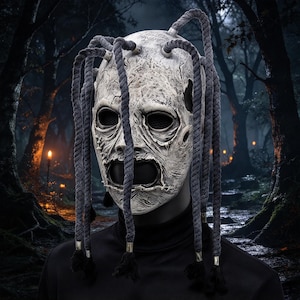 Peut inclure: Un masque intégral avec une finition blanche craquelée et vieillie, doté d'orbites noires profondes et d'une large bouche ouverte. Plusieurs cordes tressées gris foncé sont fixées sur le dessus du masque et pendent sur les côtés.