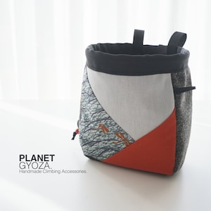 May include: A grey, white and orange climbing chalk bag with a black drawstring and a black fabric top. The bag has a geometric pattern with a dragonfly design. The text "PLANET GYOZA. Handmade Climbing Accessories." is printed on the white surface below the bag.