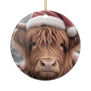 May include: A round ceramic ornament featuring a portrait of a shaggy brown Highland cow wearing a red and white Santa hat. The cow has long, textured fur, prominent horns, and a light-coloured nose. A gold string is attached to the top for hanging.