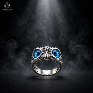 Peut inclure: Une bague argentée en forme de tête de hibou, dotée de deux yeux circulaires bleu vif et proéminents. L'anneau en métal présente des gravures complexes et texturées rappelant des plumes, offrant une apparence aviaire détaillée et réaliste.