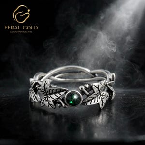 May include: A silver-toned ring featuring a band designed with sculpted ivy leaves and small berries. A single, round, dark green gemstone is set centrally within the metal foliage. The band has an intertwined, open-work structure.
