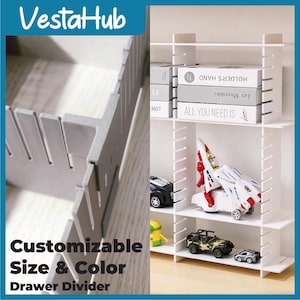 Può includere: Uno scaffale modulare bianco con pannelli verticali scanalati e ripiani orizzontali. I ripiani contengono libri intitolati Holder's Hand, Les Miserables e All You Need Is, insieme a un'astronave giocattolo bianca e due veicoli.