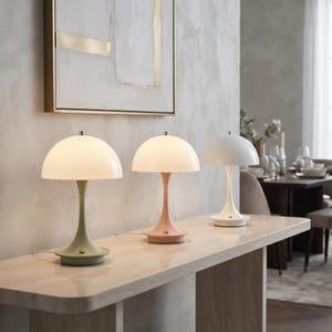 May include: Three mushroom-shaped table lamps with dome-shaped shades and slender bases. The left lamp is sage green, the middle lamp is dusty pink, and the right lamp is white. Each lamp features a small metallic switch on the circular base.