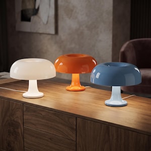 May include: Three mushroom-shaped table lamps with glossy finishes in white, orange, and blue. Each lamp features a wide, rounded dome shade and a central pedestal base, with a power cord extending from the back of each unit to provide illumination.