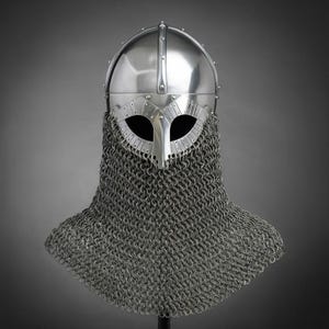 May include: A polished steel helmet featuring a rounded dome, a central vertical ridge, and a protective face guard with two eye openings. A silver-toned chainmail aventail is attached to the base, covering the neck and upper shoulder area.