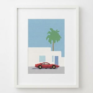 Op de afbeelding: Een ingelijst kruissteekborduurwerk met een gepixelde rode sportwagen geparkeerd voor een wit gebouw met een blauwe deur en een enkele groene palmboom tegen een lichtblauwe lucht. Het kunstwerk zit in een eenvoudige witte lijst.