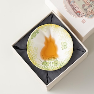 May include: A round ceramic tea cup with a scalloped edge, featuring a hand-painted design of green clover leaves and yellow foliage. The interior contains a golden-brown liquid, and the cup is presented in a white box with dark fabric.