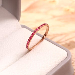 Op de afbeelding: Een dunne ring van roségoud met een doorlopende rij kleine, ronde, felroze edelstenen die langs de buitenkant zijn gezet. De ring staat rechtop en toont de gelijkmatige rangschikking van de stenen en de metalen afwerking van de band.