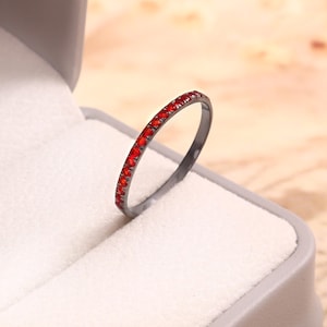 Könnte beinhalten: Ein dünner Ring aus dunkel getöntem Metall mit einer durchgehenden Reihe kleiner, leuchtend roter, runder Steine, die entlang des Bandes gefasst sind. Der Ring steht aufrecht und zeigt den Kontrast zwischen dem dunklen Metall und den roten Edelsteinen.