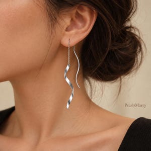 May include: A silver-toned threader earring featuring a delicate chain connected to a polished, spiralling ribbon-like metal element. The design includes a smooth, curved wire component that hangs alongside the spiral. The text PearlsMarry is visible.