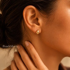 May include: A small, polished gold stud earring shaped like a teardrop. The earring features a smooth, reflective surface and is worn in the earlobe. The text 'PearlsMarry' is visible in the lower left corner of the image.