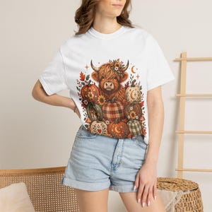 May include: A white short-sleeve t-shirt featuring a central graphic of a brown highland cow surrounded by various pumpkins, autumn leaves, sunflowers, and celestial symbols like stars and moons in shades of orange, brown, and green.