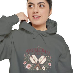 May include: A charcoal gray hooded sweatshirt featuring a graphic print that says PICKLEBALL CLUB in red letters. The design includes two crossed pickleball paddles, a ball, and various pink and white flowers with green leaves and stems.