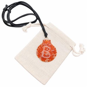May include: A necklace featuring an orange scallop shell pendant with a white letter B painted on the front. The shell hangs from a black cord, which is positioned over a small, rectangular, off-white fabric drawstring pouch.
