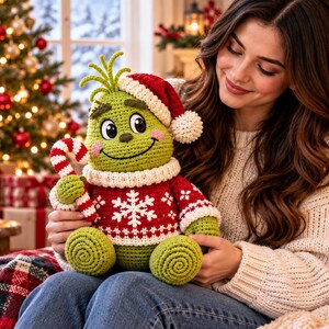May include: A green crocheted plush character with a large head, pointed tuft of hair, and a wide smile. It wears a red and white snowflake-patterned sweater, a matching Santa hat with a white pom-pom, and holds a red and white candy cane.