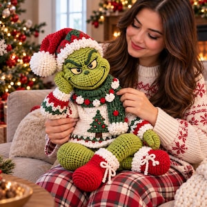Könnte beinhalten: Eine gehäkelte grüne Charakterpuppe mit mürrischem Gesichtsausdruck, die eine rot-weiße Weihnachtsmütze, einen weißen Pullover mit grünem Weihnachtsbaummuster, einen dunkelgrünen Schal mit rot-weißen Akzenten und rote Stiefel mit weißem Rand trägt.