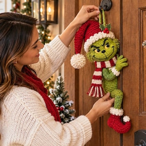 Puede incluir: Una decoración de ganchillo de una criatura verde con expresión traviesa, que lleva un gorro de Papá Noel rojo y blanco con pompón, una bufanda a rayas rojas y blancas y botas rojas con puños blancos, colgada de un gancho en una puerta.
