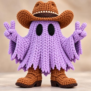 Puede incluir: Una figura de fantasma tejida a ganchillo en color morado claro, que lleva un sombrero de vaquero marrón y botas de vaquero a juego. El fantasma tiene dos ojos ovalados negros y mantiene ambos brazos en alto haciendo el signo de la paz.