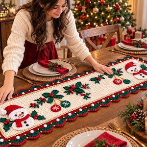 Puede incluir: Un camino de mesa rectangular de ganchillo color crema con dos figuras de muñecos de nieve con gorros y bufandas rojas, decorado con hojas de acebo verdes, bayas rojas y adornos colgantes, con un borde festoneado en rojo y verde.