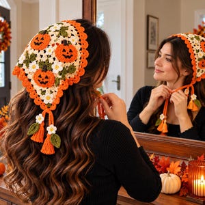 Puede incluir: Un pañuelo triangular de ganchillo con un patrón de calabazas naranjas, margaritas blancas con centro amarillo y hojas verdes. Los bordes están rematados con un ribete naranja festoneado y dos borlas naranjas colgantes.