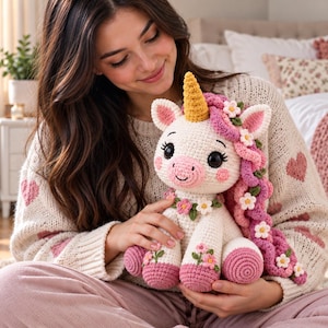 Op de afbeelding: Een gehaakte eenhoorn knuffel met een crèmekleurig lichaam, een goudgele hoorn en een lange, krullende roze manen versierd met kleine witte en gele bloemetjes. De eenhoorn heeft roze hoeven, roze wangen en bloemmotieven op de voorpoten.