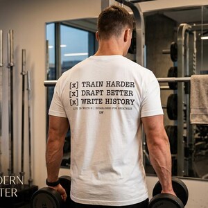 May include: A white short-sleeved t-shirt featuring black text on the back that reads [x] TRAIN HARDER [x] DRAFT BETTER [x] WRITE HISTORY, with smaller text below stating LIFE IS WRITE | ESTABLISHED FOR GREATNESS | 136.