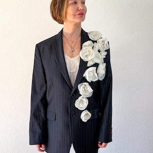 Puede incluir: Un blazer gris oscuro de rayas diplomáticas con un grupo de flores de tela blanca sujetas en la solapa izquierda y el pecho. El blazer tiene cuello de solapa, dos botones frontales y dos bolsillos con solapa, sobre una camiseta blanca.