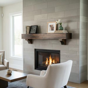 May include: A dark brown wooden fireplace mantel mounted on a light gray tiled wall above a gas fireplace with visible flames. The mantel holds a black square clock, two books, a framed picture, and a small green vase with a white flower.