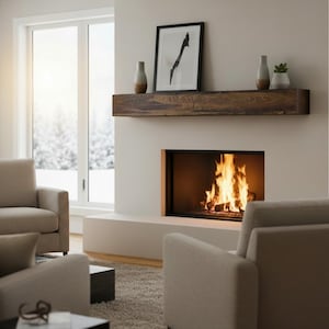 May include: A dark brown wooden fireplace mantel mounted on a light wall above a burning fireplace. The mantel holds two grey vases, a framed black and white abstract print, and a small potted succulent plant in a white ceramic container.