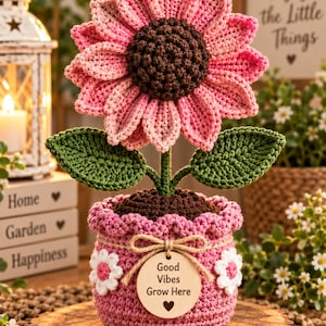 Könnte beinhalten: Eine gehäkelte rosa Sonnenblume mit dunkelbrauner Mitte und zwei grünen Blättern in einem strukturierten rosa Häkeltopf. Der Topf ist mit zwei weißen Häkelblumen und einem runden Holzanhänger mit der Aufschrift Good Vibes Grow Here verziert.