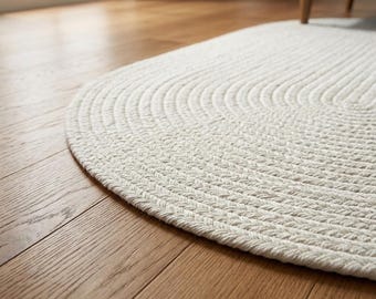 Könnte beinhalten: Ein ovaler Teppich aus dickem, geflochtenem cremeweißem Seil. Der Teppich weist ein durchgehendes, konzentrisches Muster aus eng gewebten Fasern auf, die eine texturierte, gerippte Oberfläche über den gesamten Bodenbelag erzeugen.