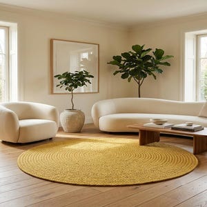 May include: A large, round, mustard yellow braided area rug with a concentric circular pattern. The rug is placed on a light-colored wooden floor in a room featuring a cream-colored curved sofa, a matching armchair, and two potted plants.