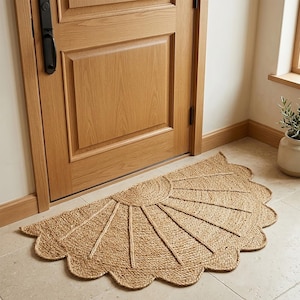 May include: A semi-circular natural fiber doormat featuring a sunburst pattern with radiating lines and a scalloped edge. The mat is woven from tan, textured material and is positioned on a light-colored tiled floor in front of a wooden door.