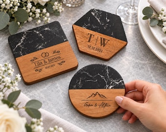 May include: Three coasters made of black marble and wood. One square coaster reads Lila & James 03.07.2023, one hexagonal coaster reads T | W 12.12.2022, and one circular coaster held by a hand features a mountain graphic and Golem & Althea.