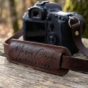 May include: A dark brown leather camera strap featuring a padded shoulder section with the embossed text Mason Clarke PHOTOGRAPHY in a combination of script and sans-serif fonts, attached to a black digital camera body with a metal ring.